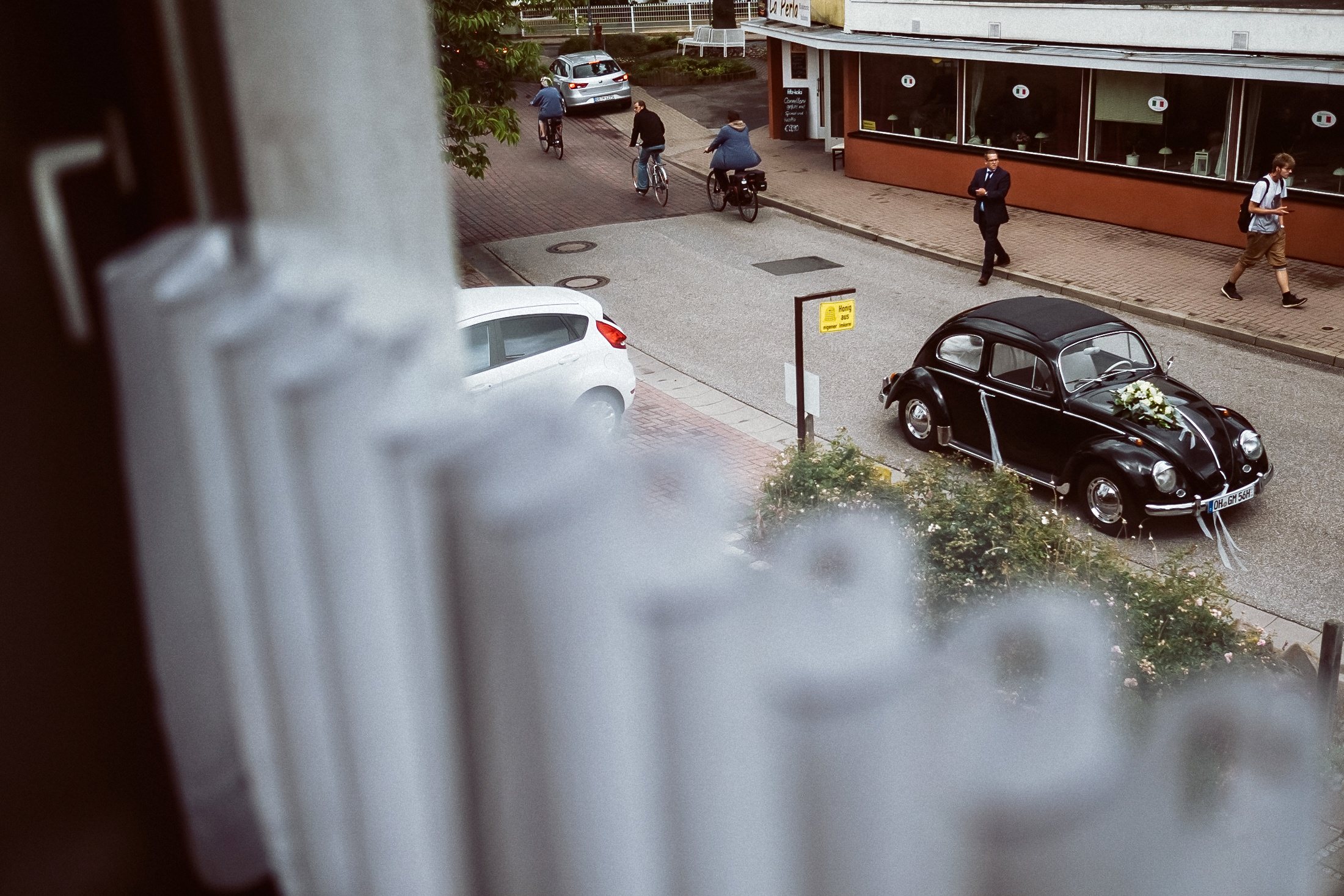 Schwarzer VW Käfer mit Brautstrauß auf der Ortsstraße, von oben durch die Gardine fotografiert.